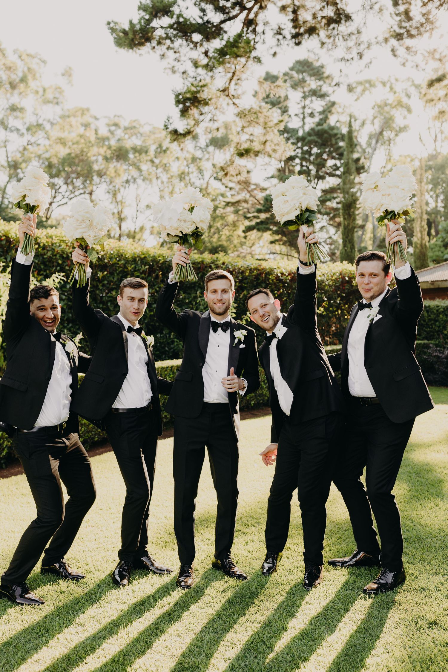 Wedding party in black tuxedos stands in line making playful poses outdoors.