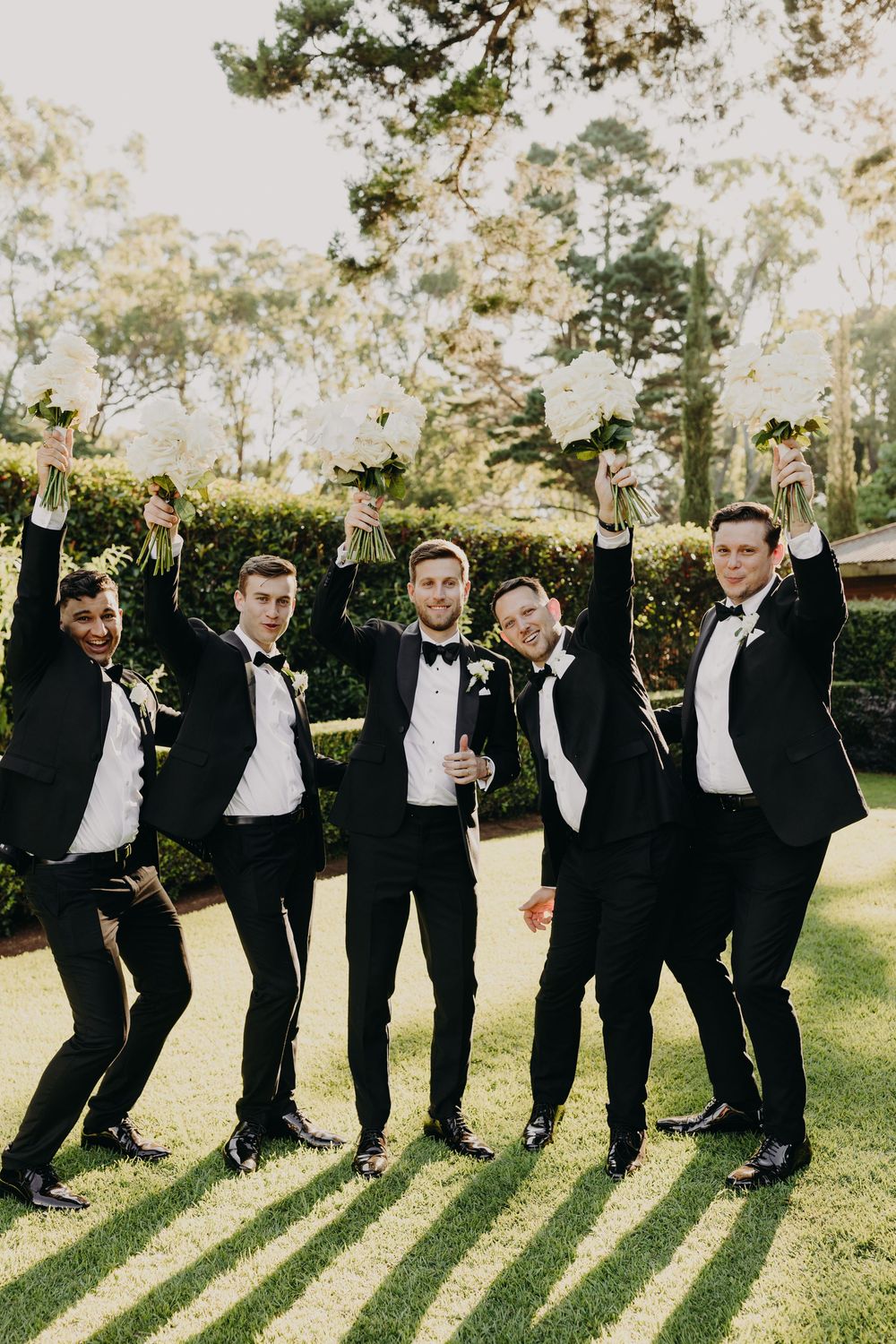 Wedding party in black tuxedos stands in line making playful poses outdoors.