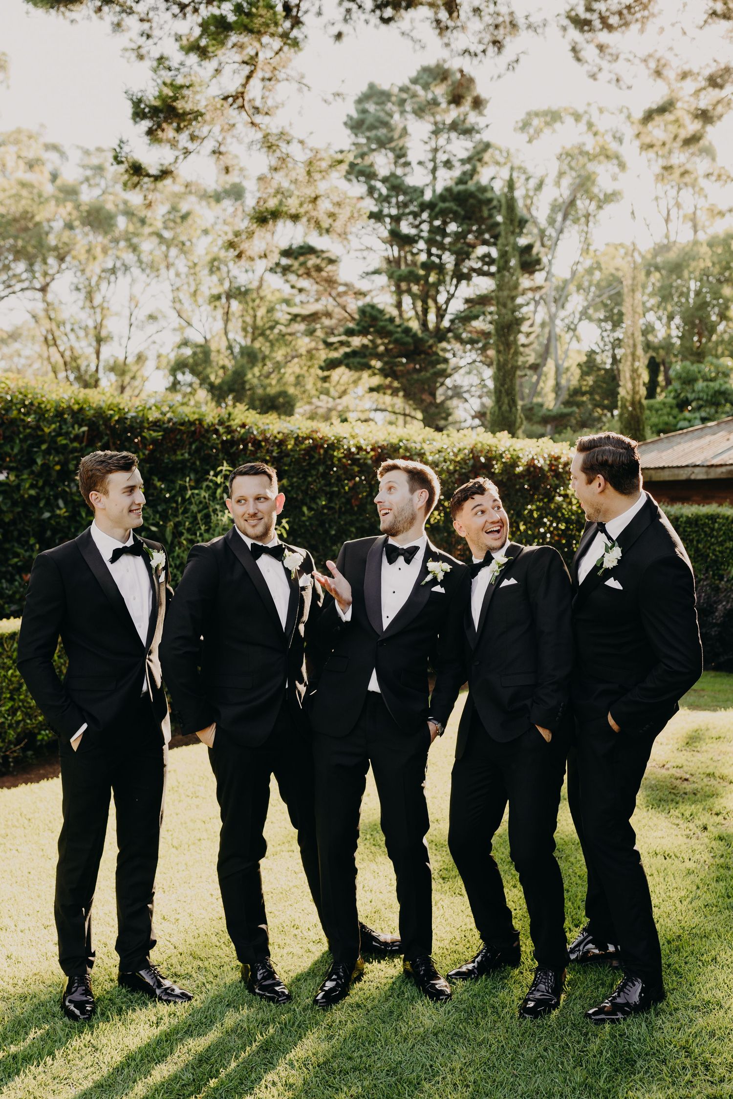 Groomsmen lined up outdoors share laughs in classic black tuxedos.