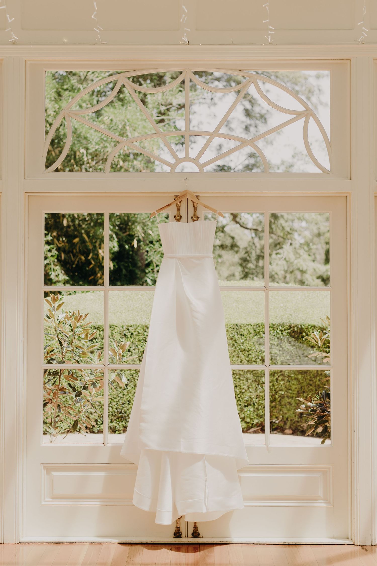 White wedding dresses hang elegantly in sunlit windows of a bridal preparation room.