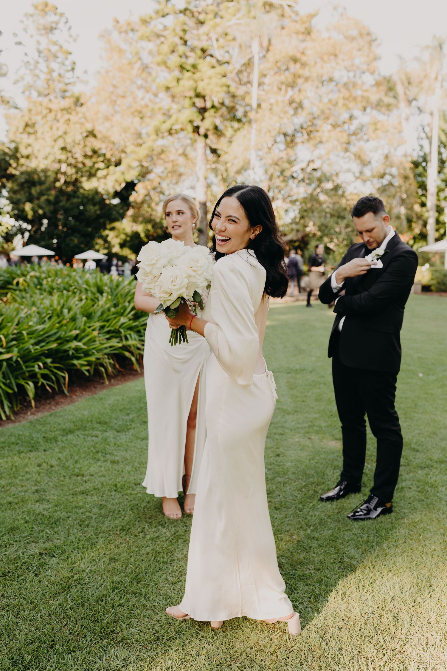 Wedding party in white dresses and black tuxedos pose casually on green lawn.