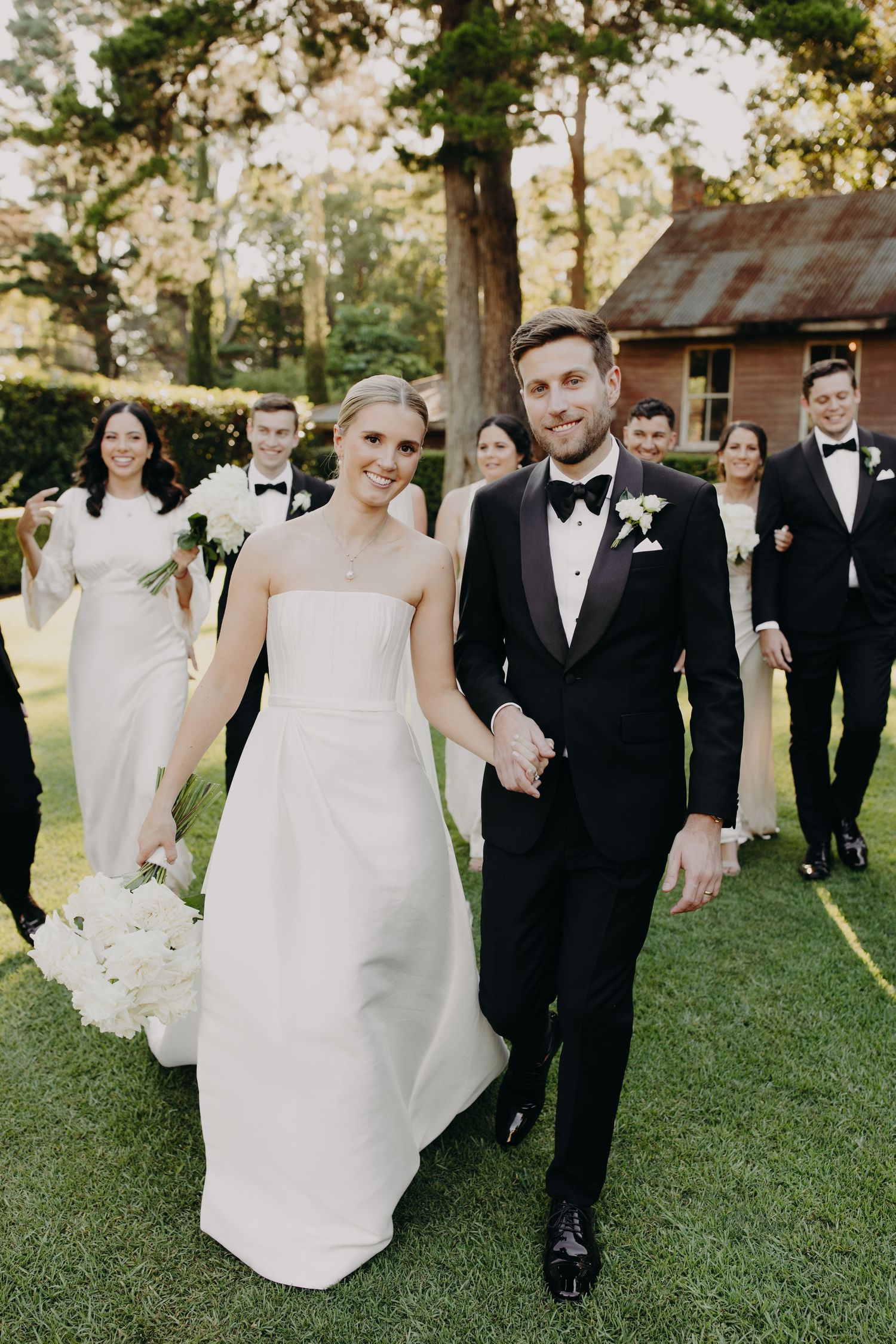 Bridal party in white dresses and black tuxedos walks together on manicured lawn.