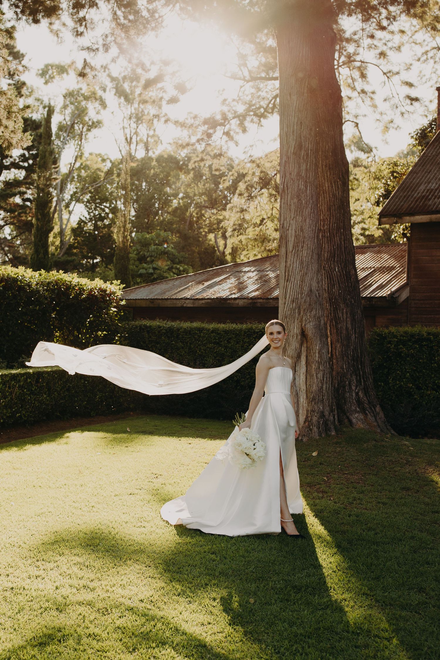 Flowing white wedding dress catches sunlight against rustic outdoor backdrop.