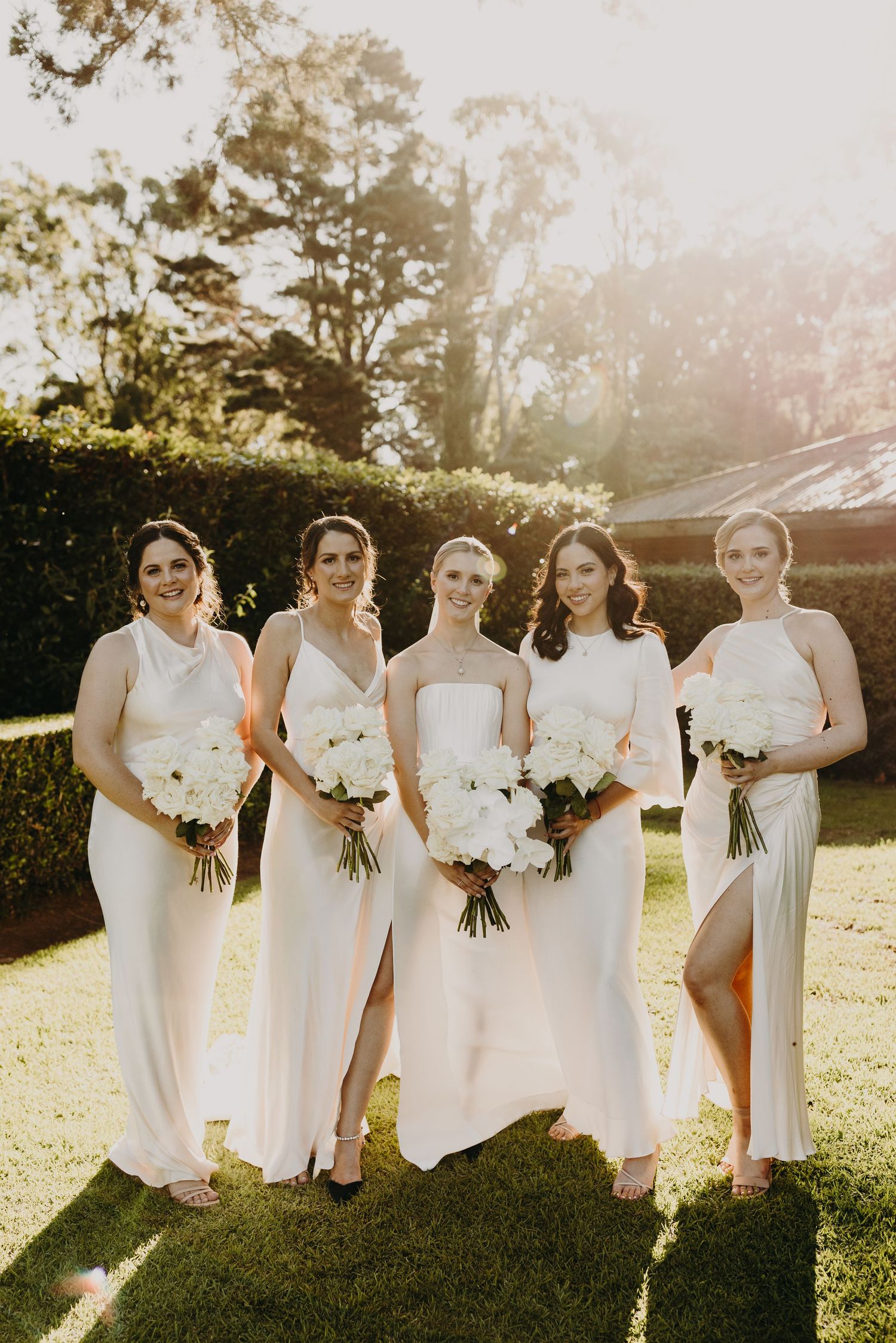 Bridesmaids in white dresses hold matching bouquets during golden hour photo.