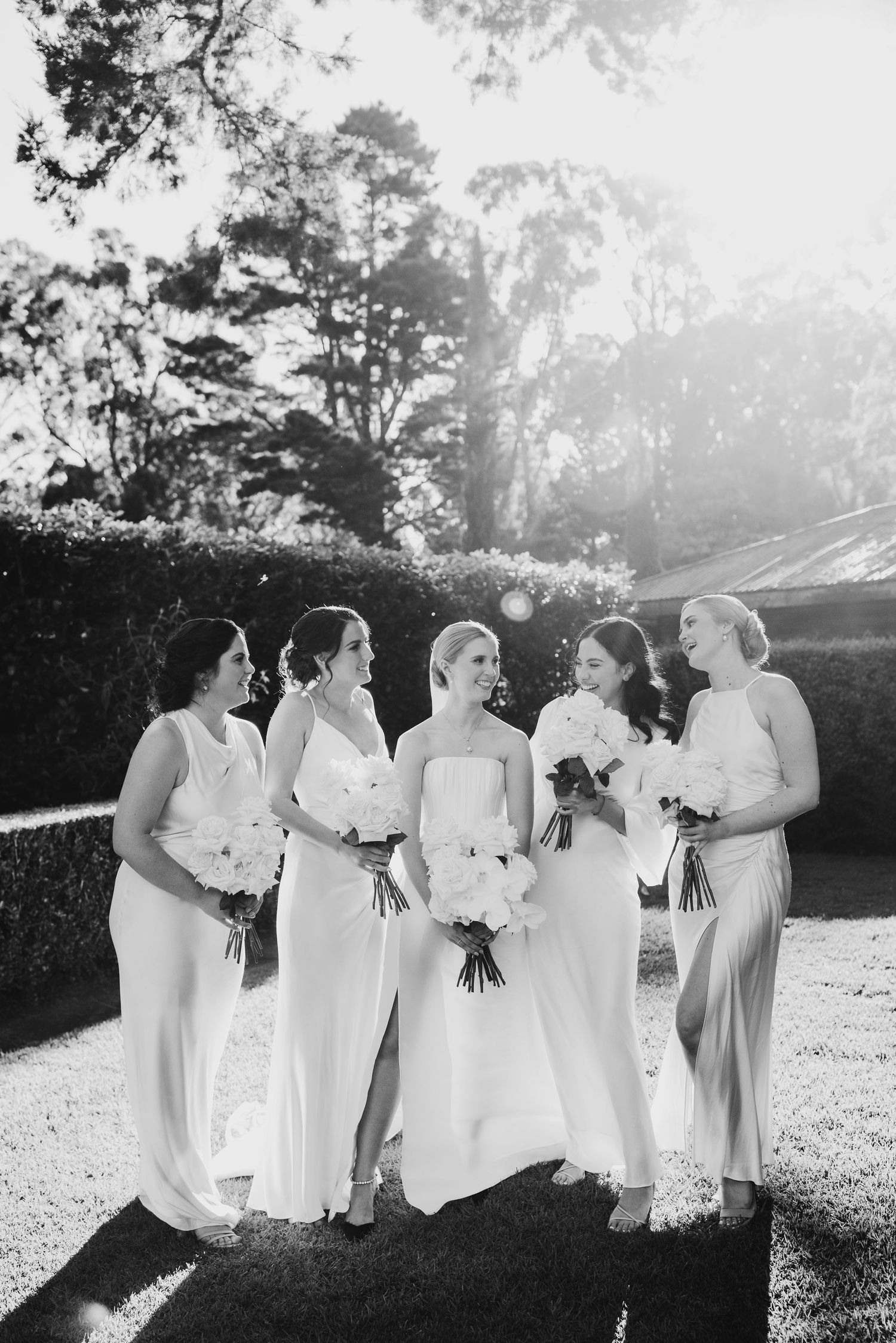 A group of bridesmaids in white dresses stand together outdoors during a wedding photoshoot.
