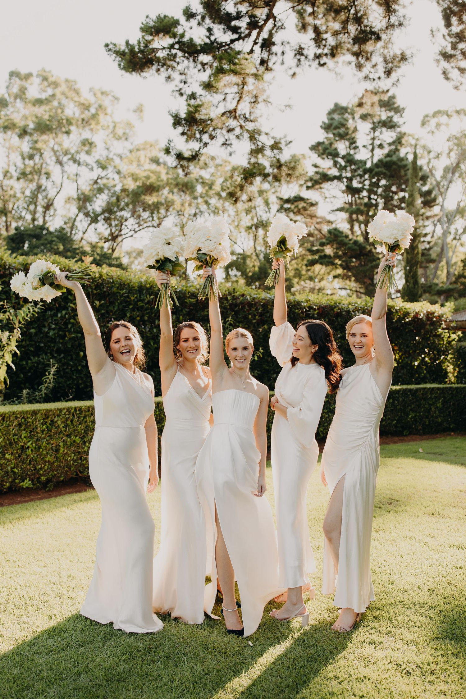 Bridesmaids in flowing white dresses celebrate joyfully on a sunlit lawn.