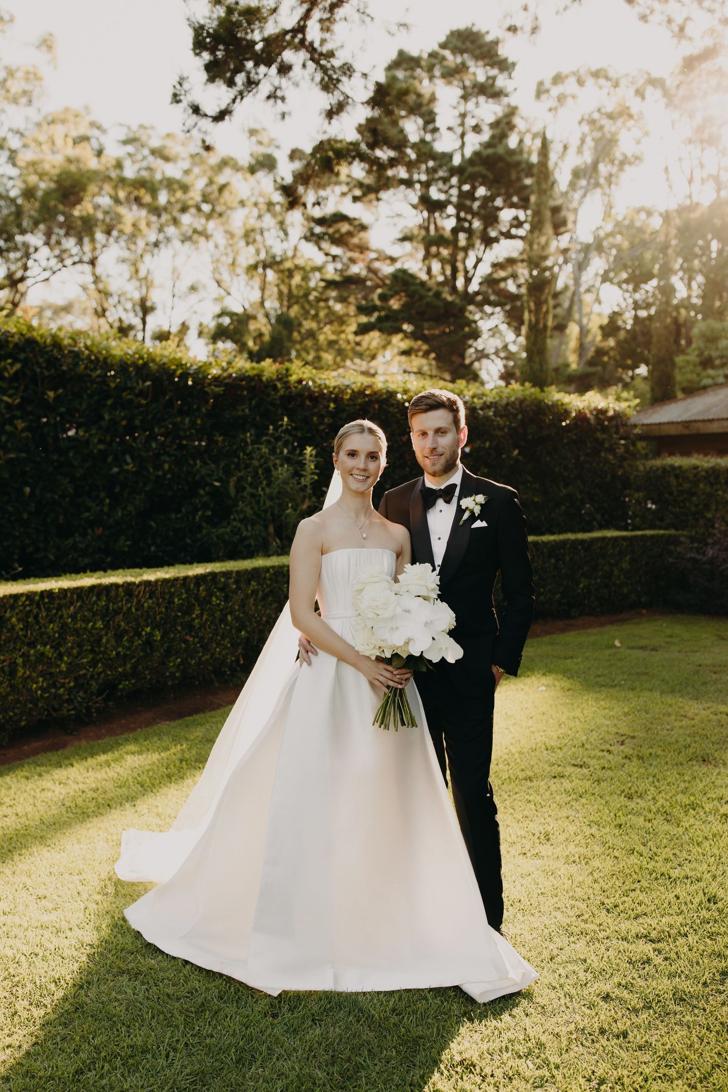 A couple poses for romantic wedding photos on a sunlit lawn with hedge backdrop.