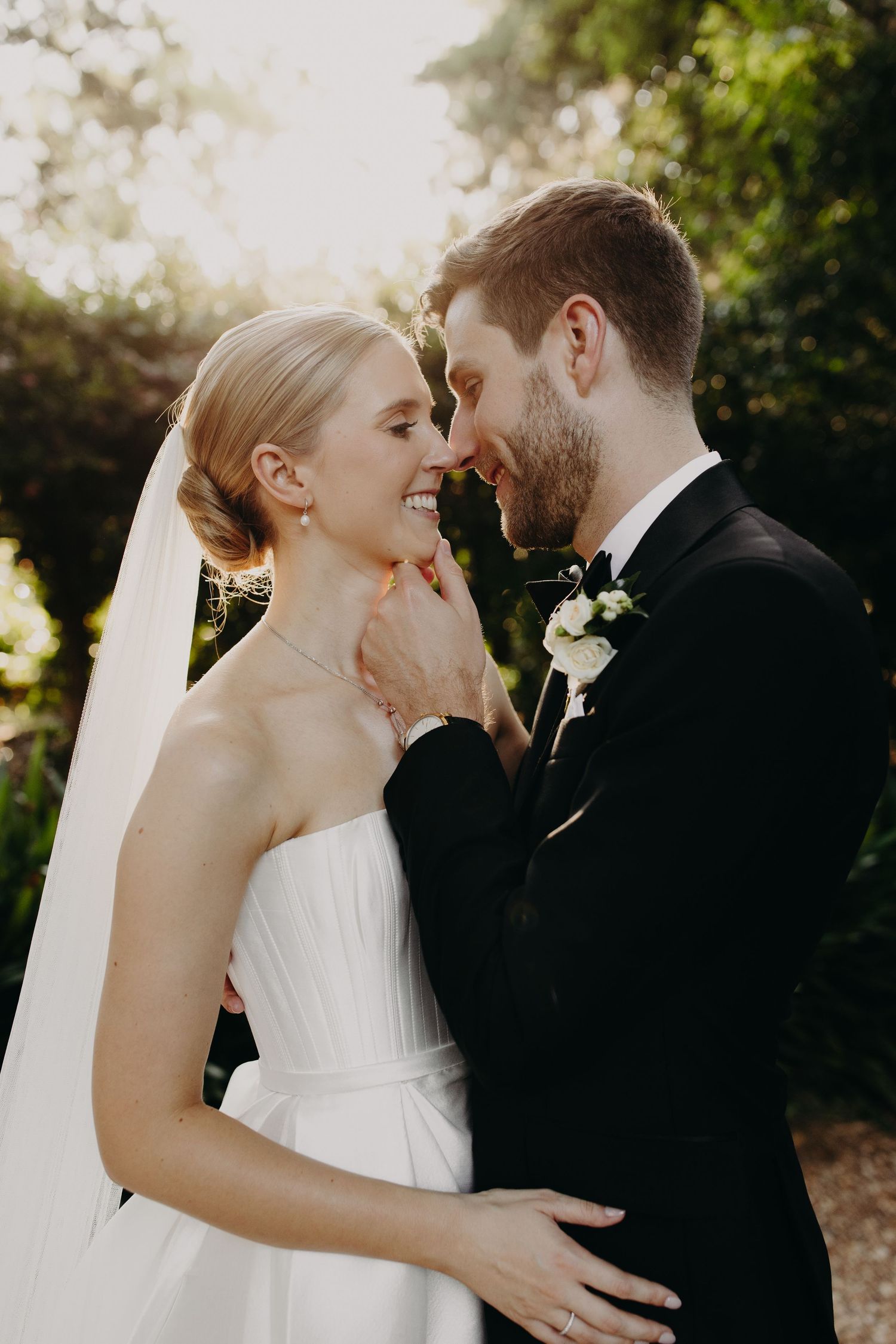 Newlyweds embrace tenderly in a sunlit outdoor setting with the bride wearing a flowing white veil.