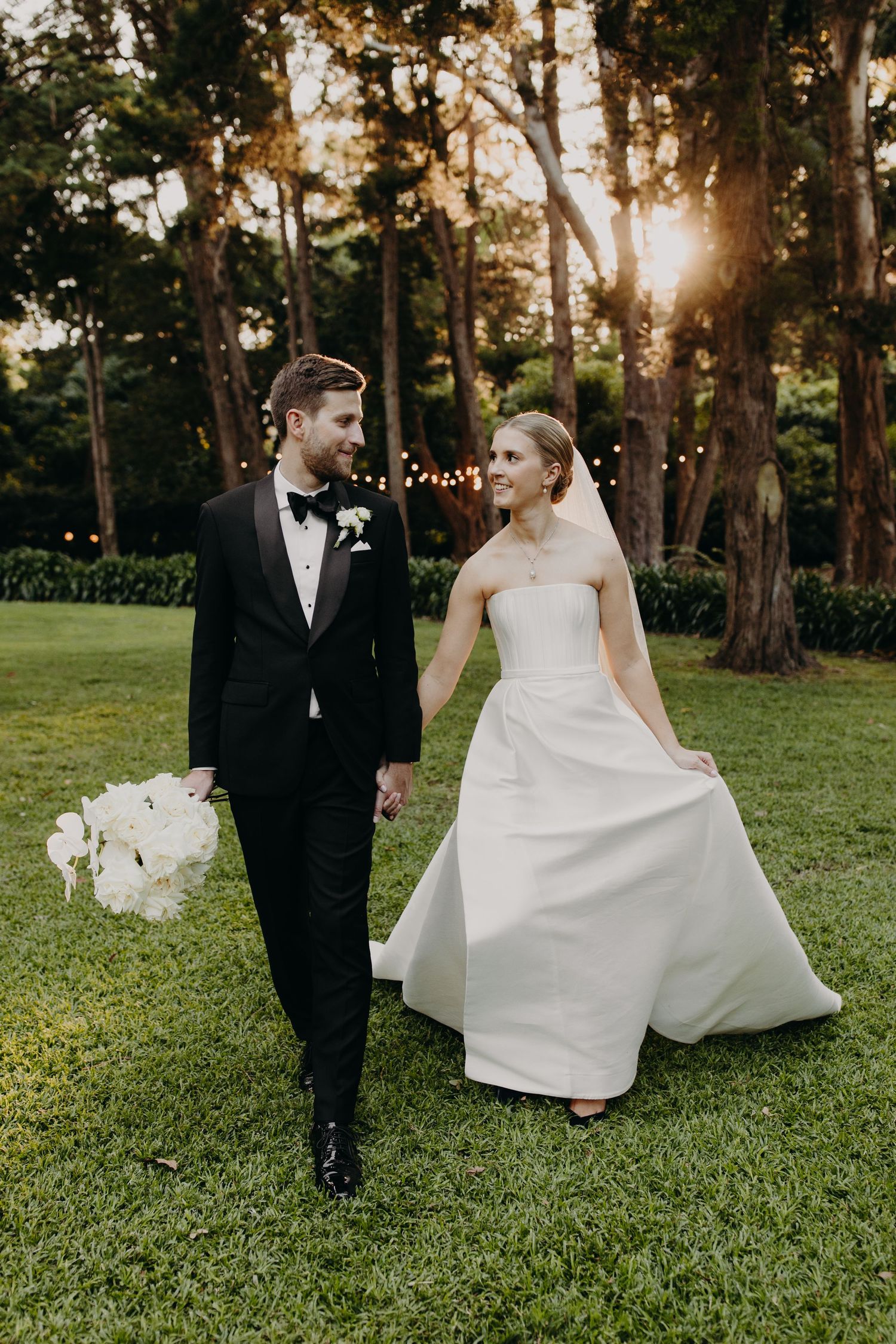 Wedding couple walks playfully across a sunlit lawn during their outdoor photo session.