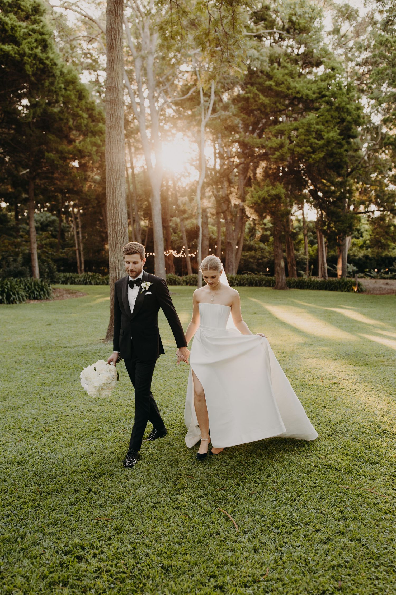 Bride in elegant white dress and groom explore garden grounds during golden hour photography session.
