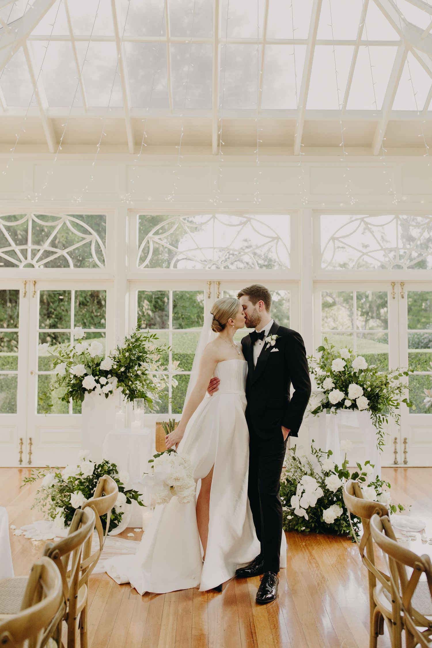 Elegant wedding reception in a bright conservatory with white floral arrangements and wooden cross-back chairs.