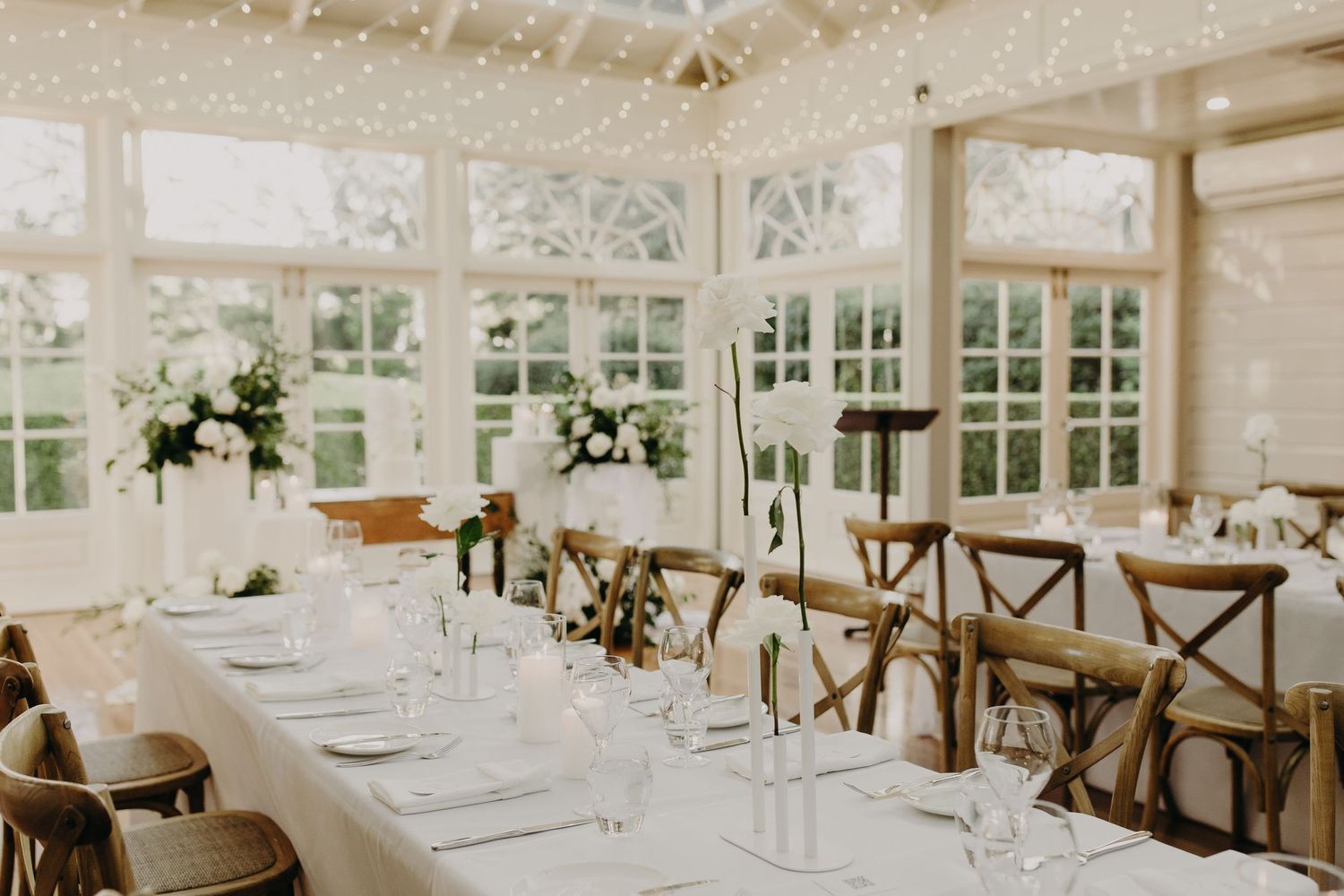 Light-filled wedding venue with long tables set for a reception in a white conservatory space.