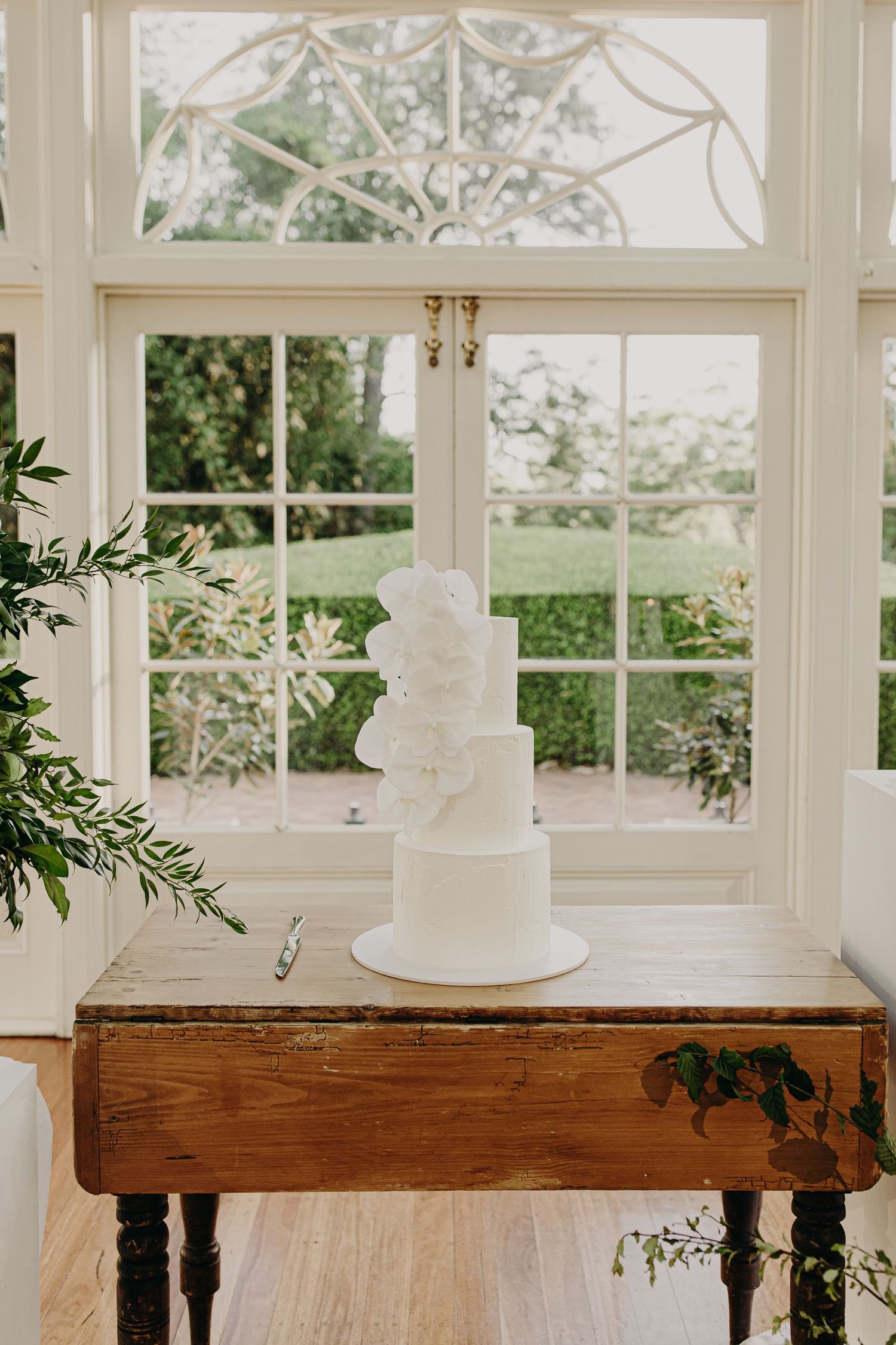 White wedding cake displayed on rustic wooden table with greenery accents in sunlit conservatory.
