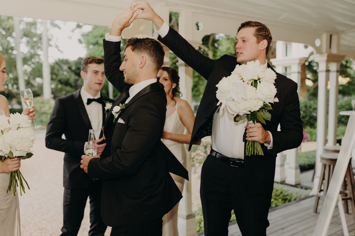 Wedding party in formal attire gathered in bright indoor venue with white floral decorations.