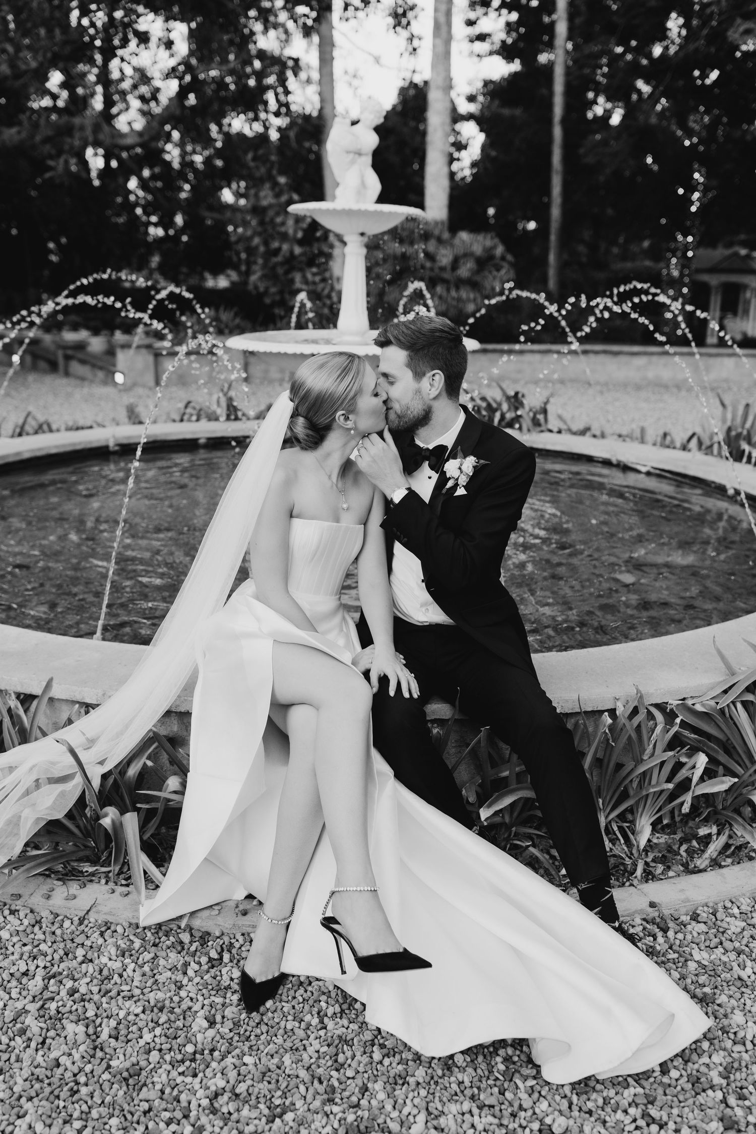 Black and white photo of newlyweds sitting together by decorative fountain.