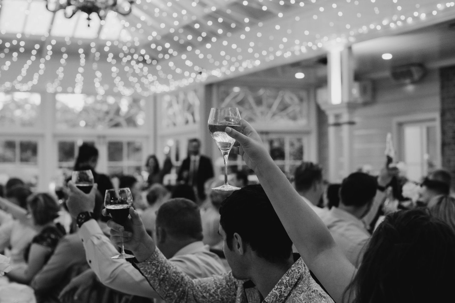 Wedding guests raise their glasses in celebration under twinkling lights at an indoor reception venue.