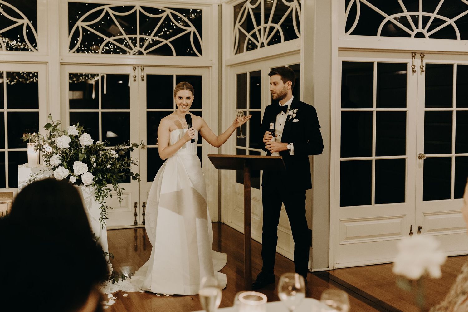 Wedding celebration with speeches in an elegant room with ornate windows and floral decor.