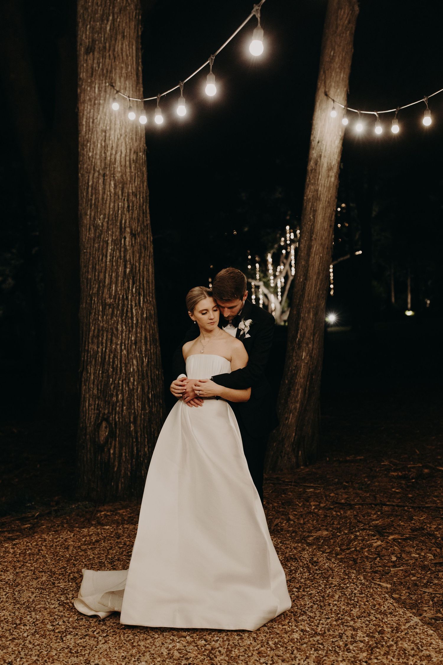 Couple dancing under string lights in a wooded outdoor setting at night.