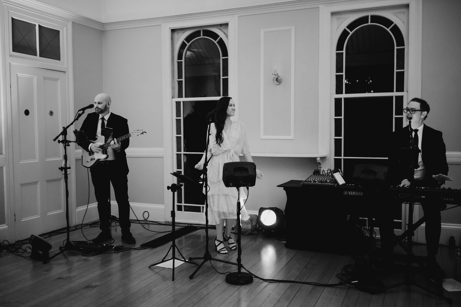 Black and white photo of musicians performing in an elegant room with arched windows.