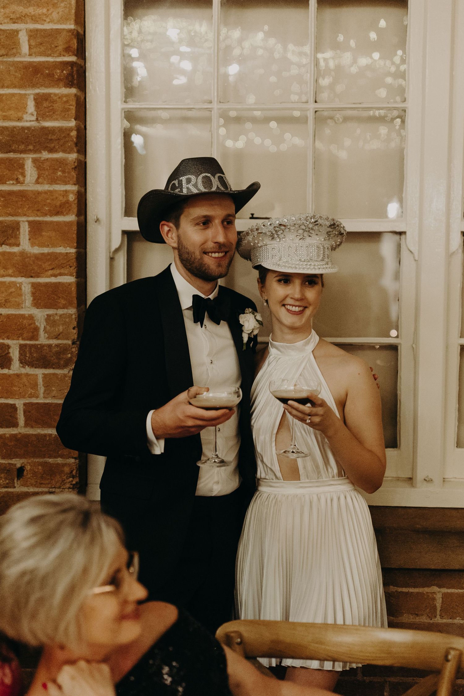 Wedding party guests enjoying drinks in a rustic brick-walled venue with string lights.