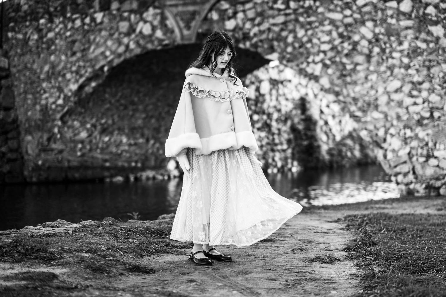 Black and white image of a young girl watching her dress flow in the breeze near a bridge