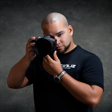 Professional photographer holding a DSLR camera, wearing a black t-shirt against a studio backdrop.
