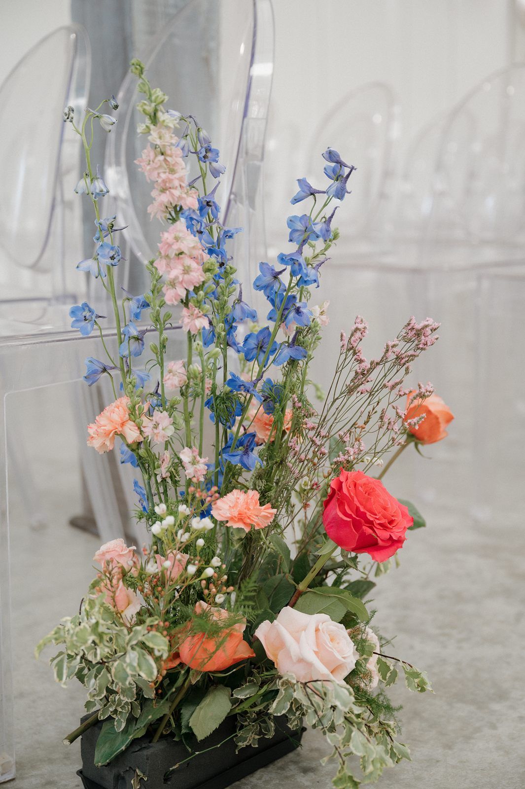 A delicate floral arrangement with blue delphiniums, peach and coral roses, and soft greenery in a dark container.