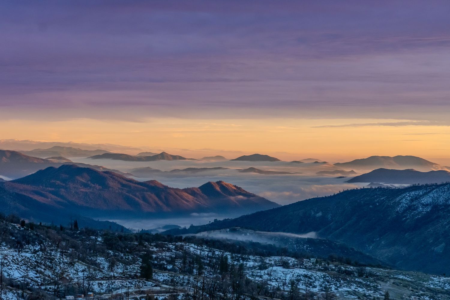 Scenic panoramic view of snowy mountain ranges with dramatic purple and orange sunset skies above misty valleys.