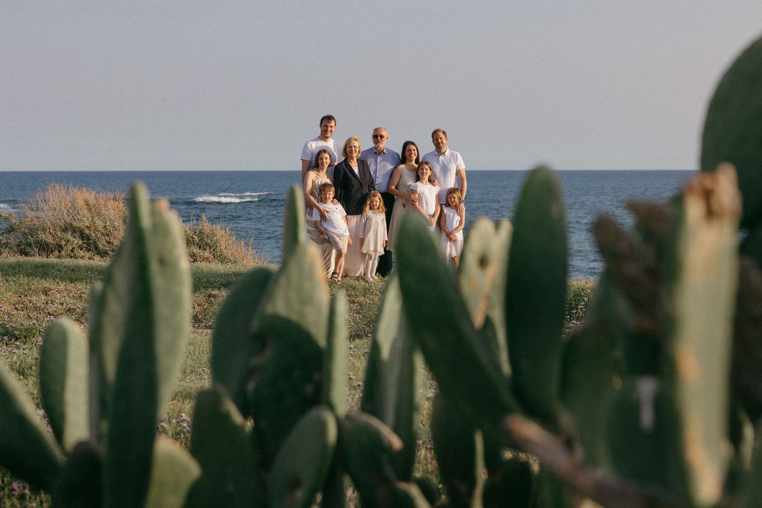 Family group portrait in white outfits on coastal cliffside with cacti foreground and blue sea backdrop.