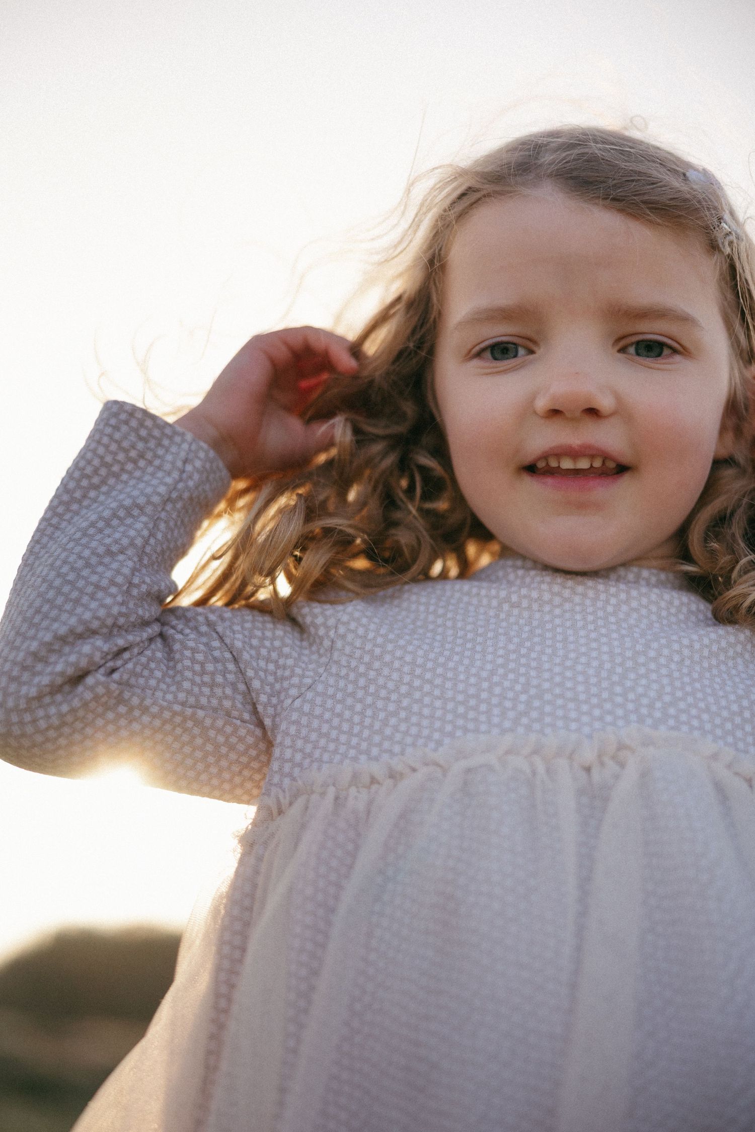 A young girl with curly hair in a white dotted dress smiles outdoors in golden sunset light.