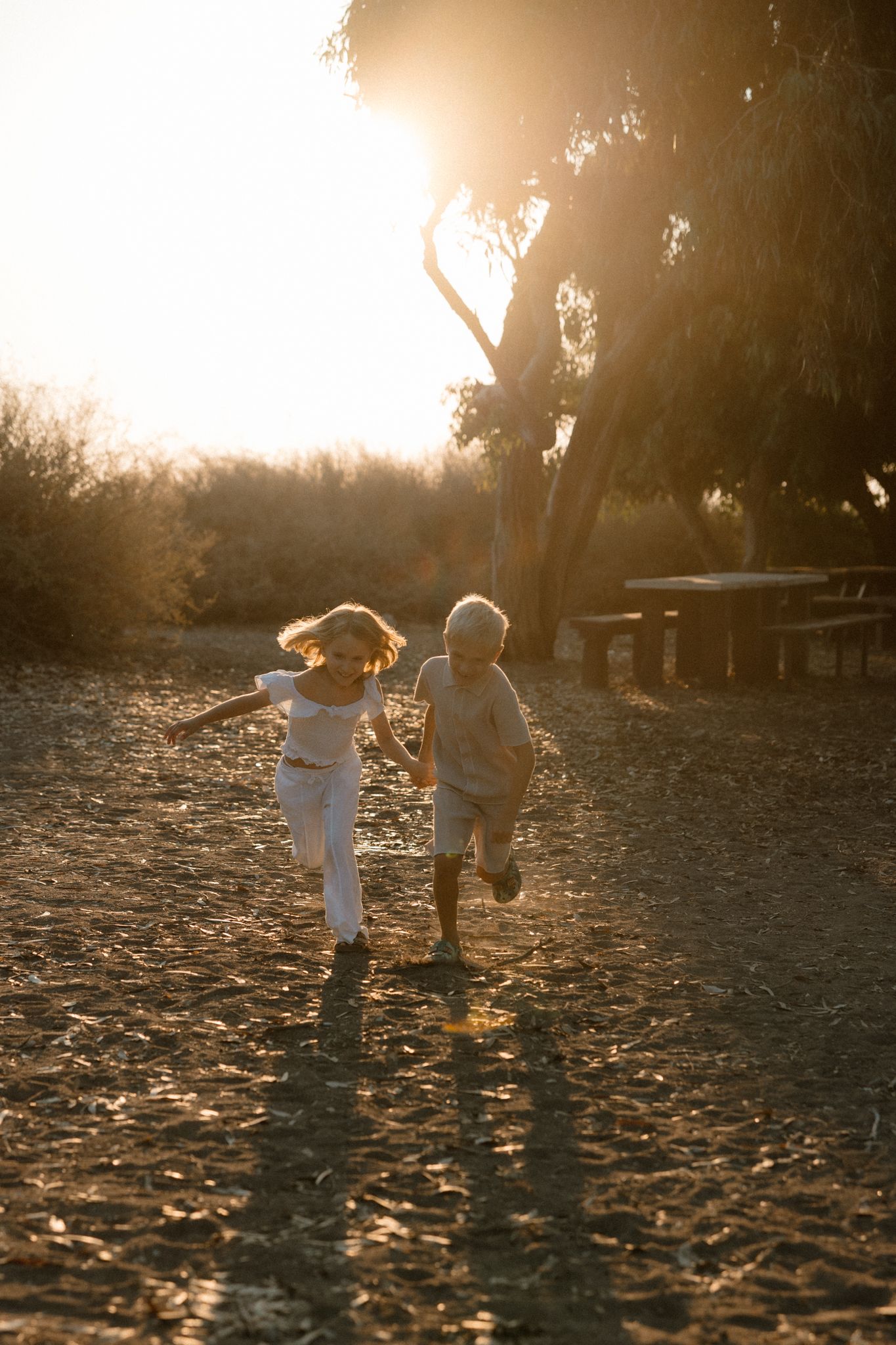 Two children running together on sandy ground during golden sunset hour near trees.