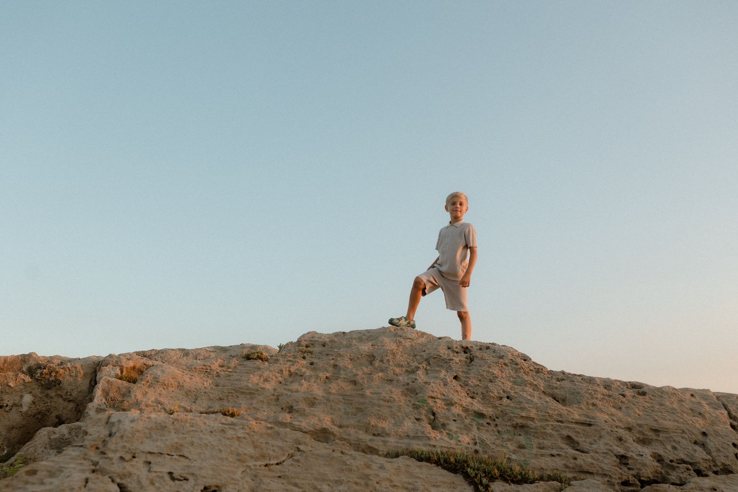 A young boy stands confidently on rocky terrain against a clear blue sky at sunset.