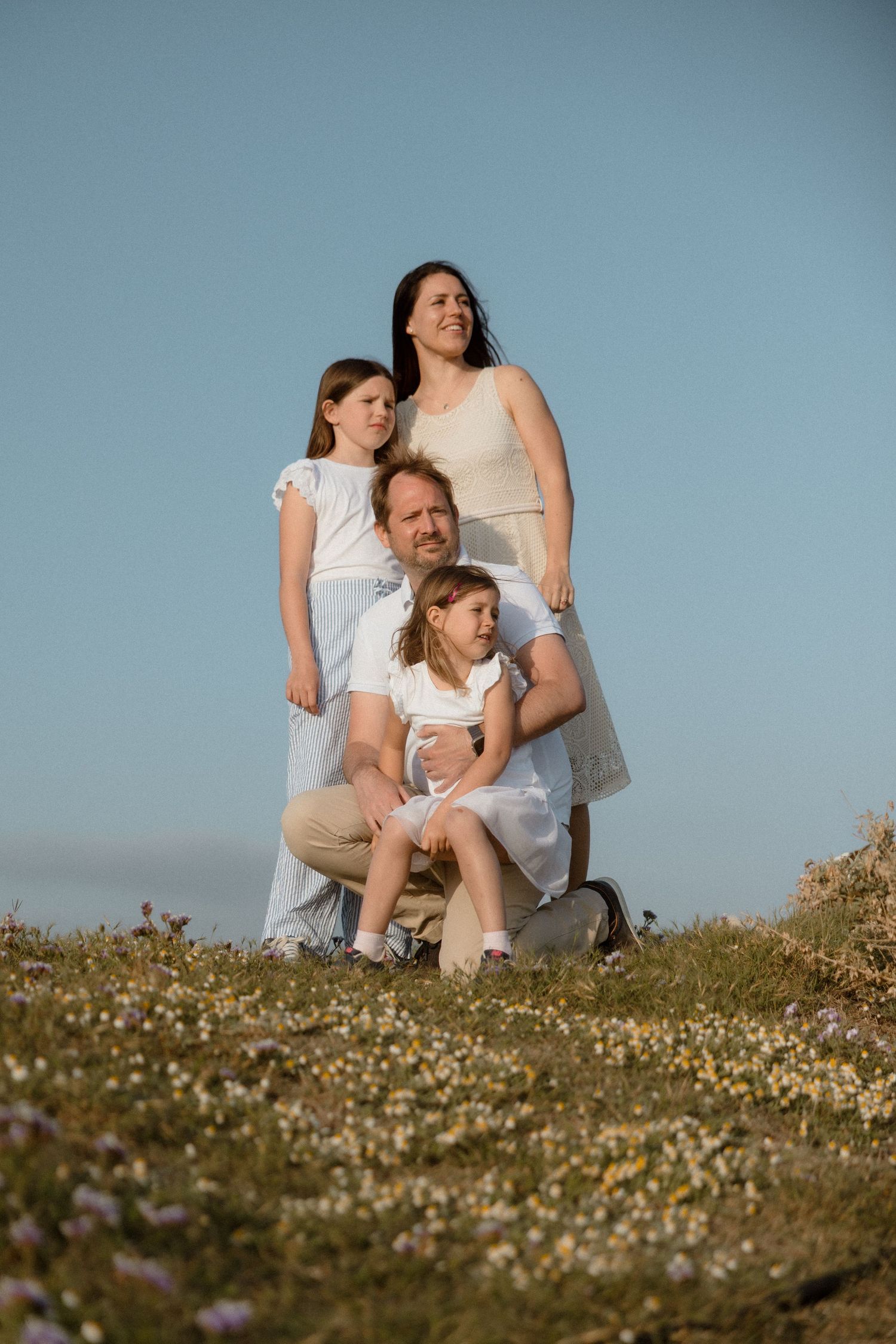 Family of four posing outdoors in white outfits on a flower meadow against a clear blue sky.