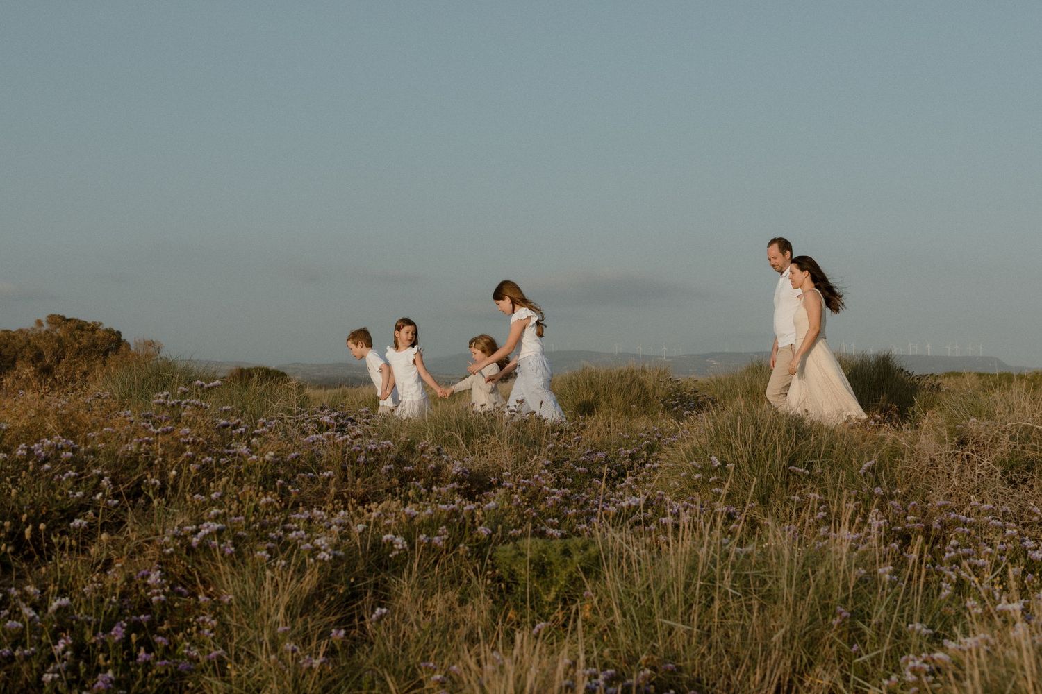Family in white attire walking through a wildflower field at golden hour with a serene coastal backdrop.