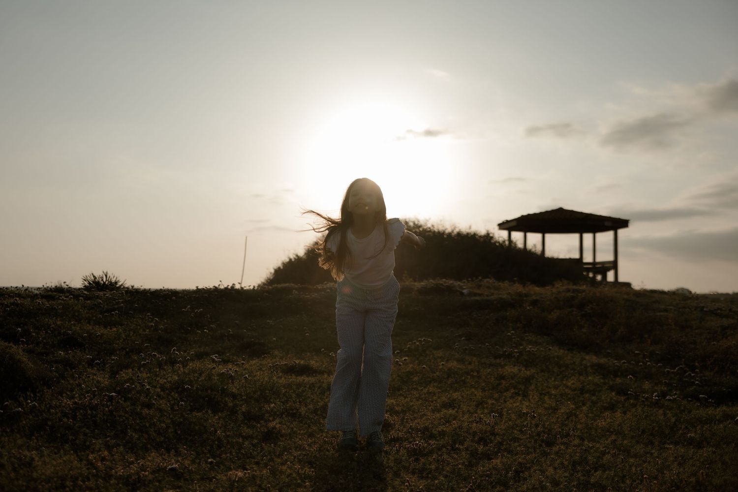 Woman standing in a sunlit field at golden hour with a gazebo and glowing sky in the background.