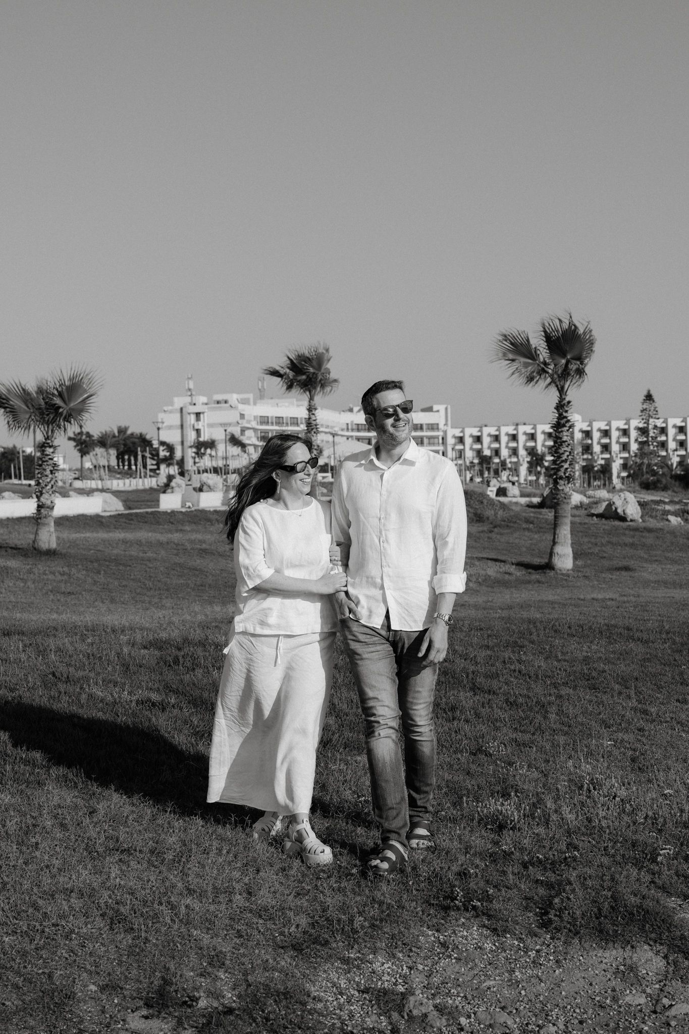 Black and white photo of a couple in white outfits and sunglasses standing on grass with palm trees and a resort.