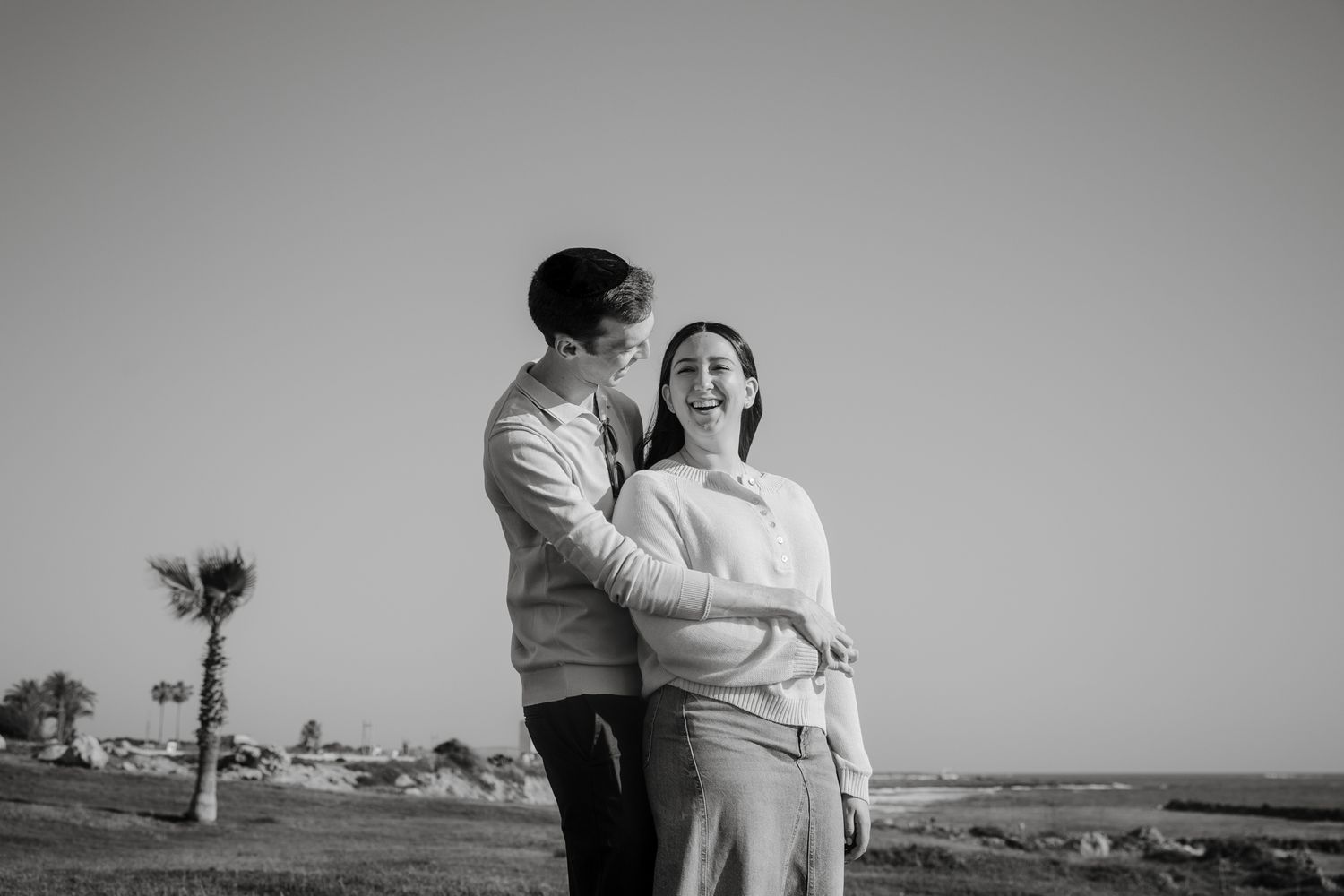 Black and white photo of a happy couple embracing outdoors near a coastal landscape with a palm tree in the background.