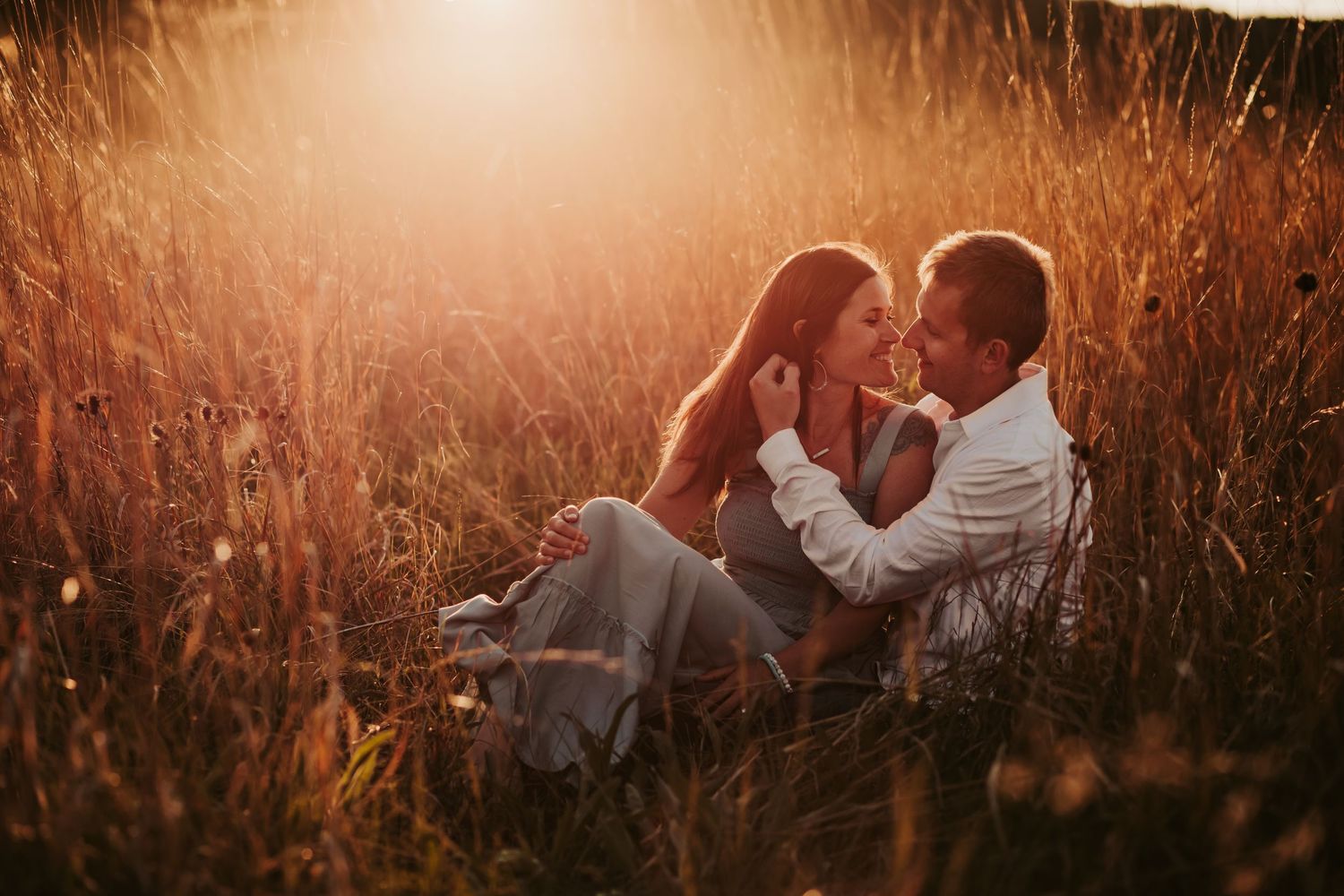 Couple embracing in tall grass field during golden hour sunset.