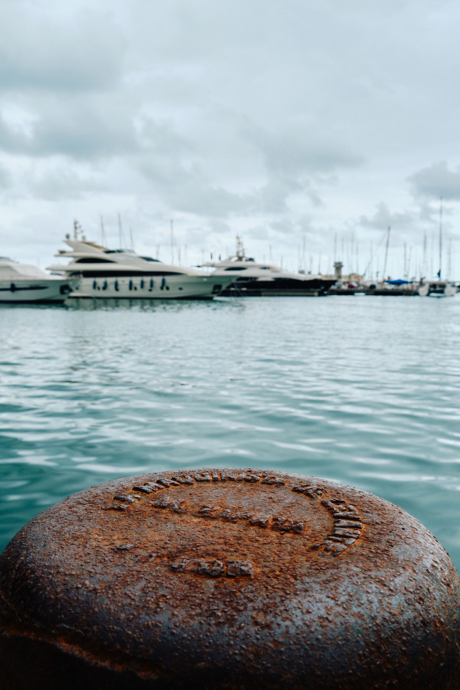 Luxury yachts docked in a marina with turquoise waters and a rusty mooring bollard in the foreground.