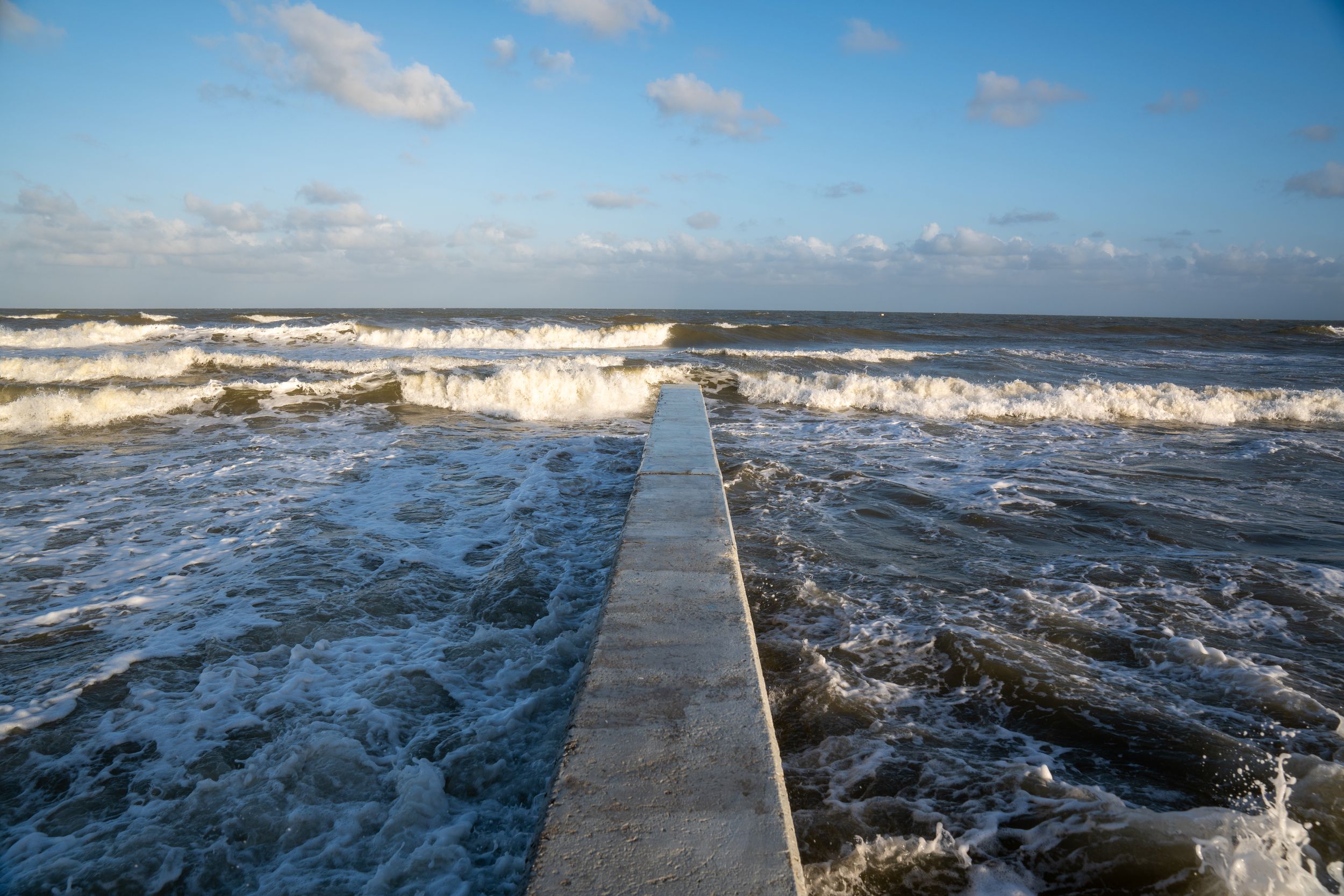 Dramatic waves crash along a concrete pier extending into the blue ocean under cloudy skies.