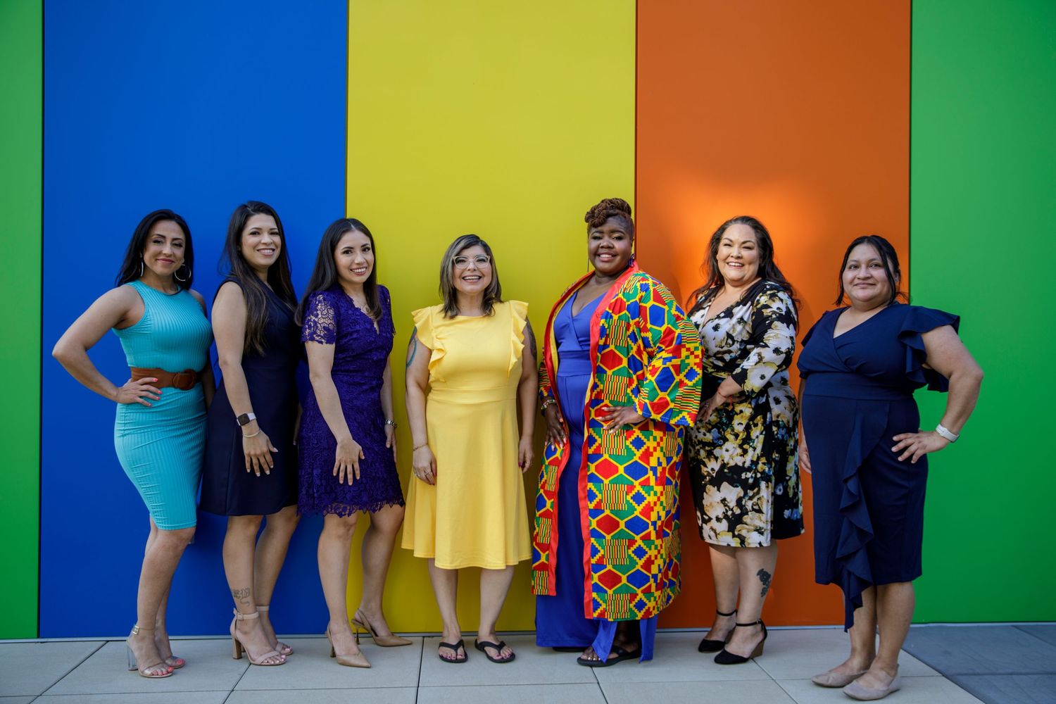 Group photo of people wearing colorful dresses against vibrant striped wall backdrop.