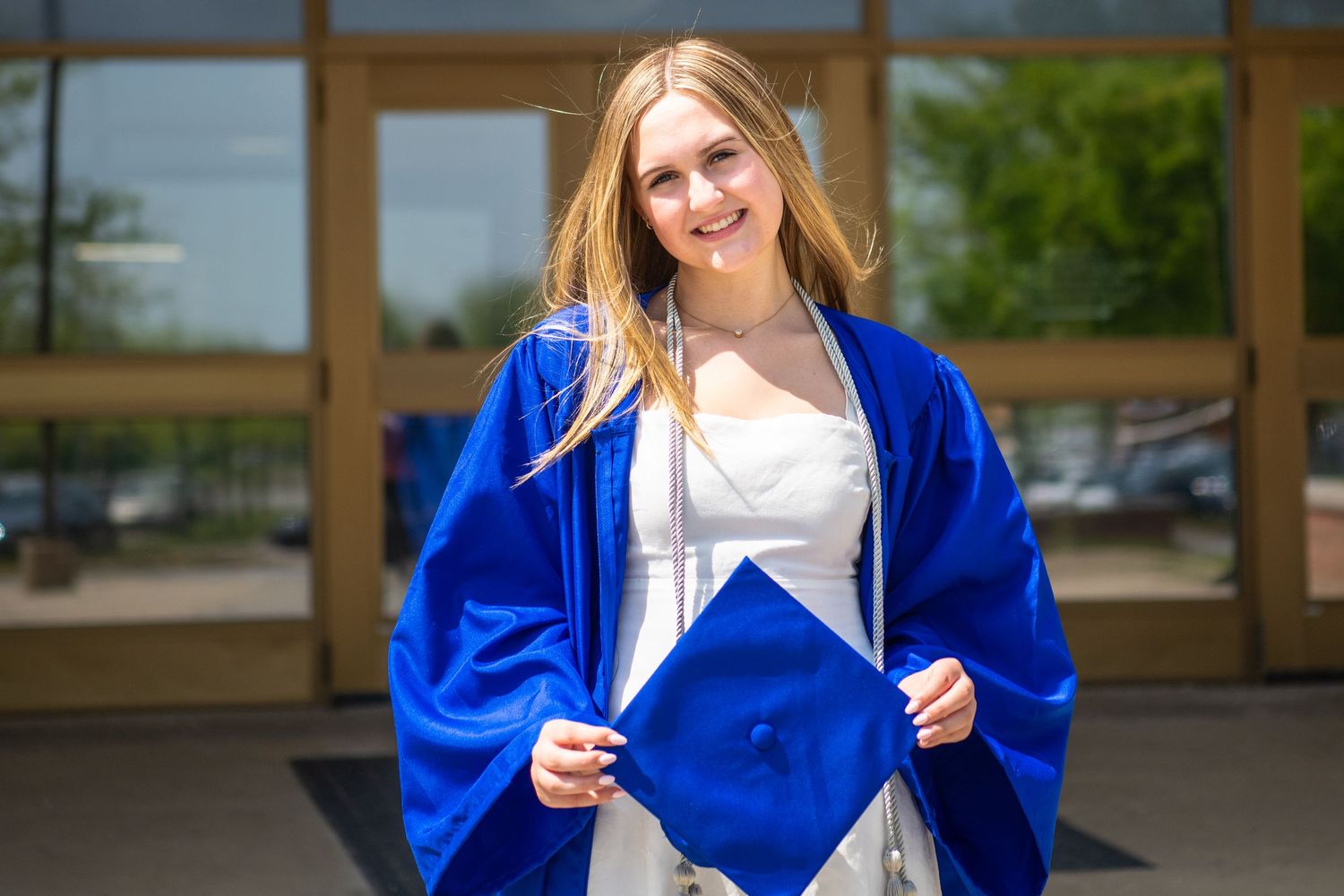 Graduate in bright royal blue graduation gown and white dress stands outside holding cap on commencement day.
