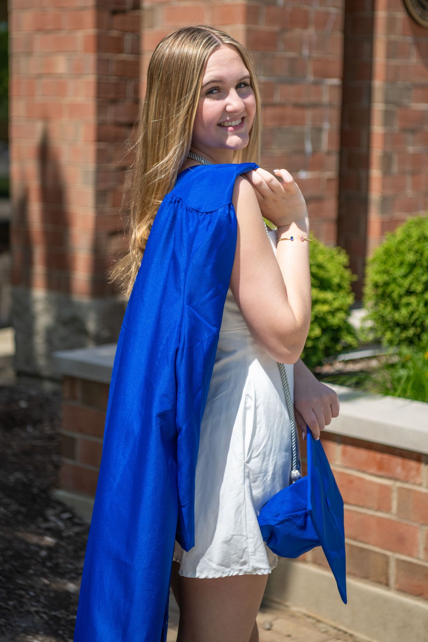A graduate in a royal blue graduation gown poses outdoors against a brick building on a sunny day.