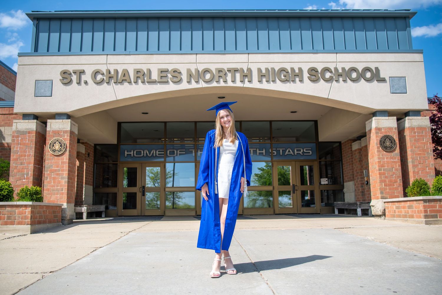 Graduate in blue cap and gown stands in front of St. Charles North High School entrance on sunny day.