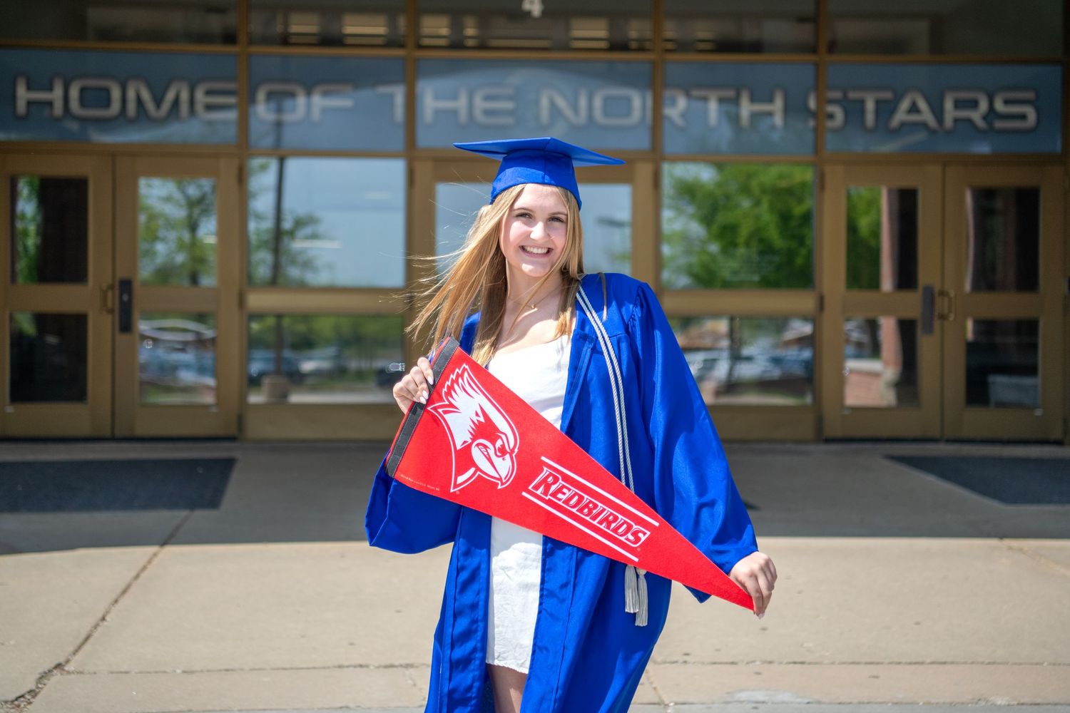 Graduate in blue cap and gown holds a red pennant while standing in front of North Stars building entrance.