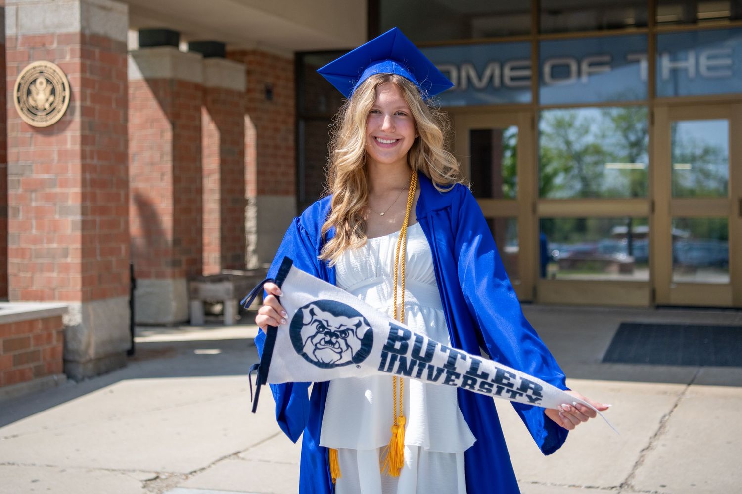 Graduate in blue cap and gown holds up Penn State University banner while smiling outside campus building.
