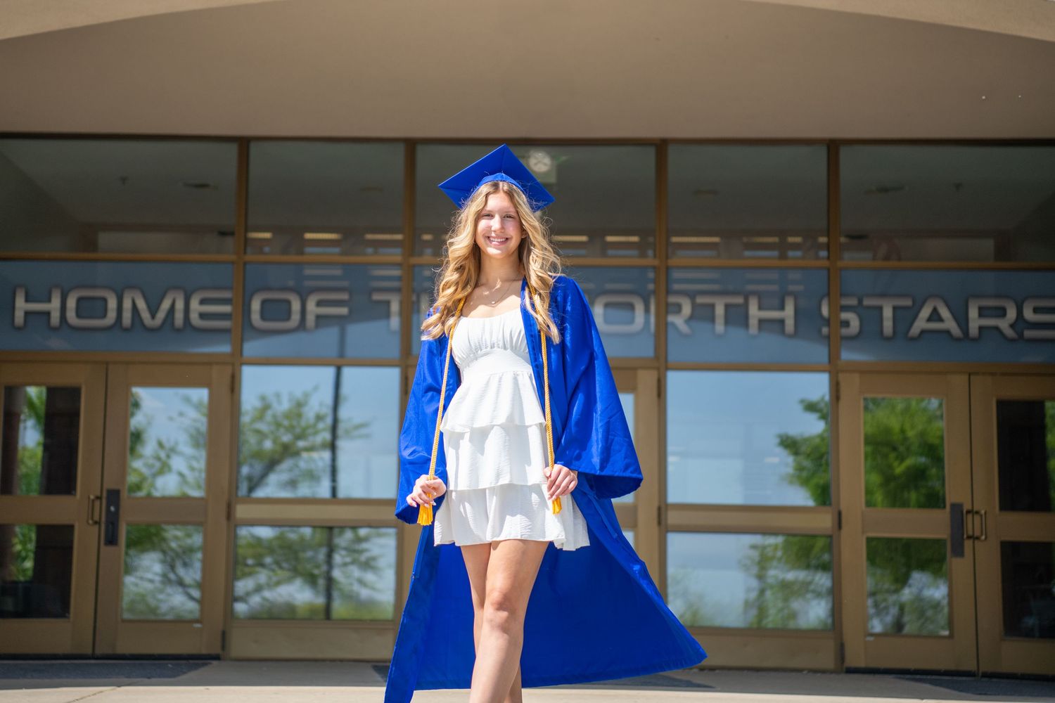 Graduate in blue cap and gown stands proudly in front of North Stars school entrance during graduation day.