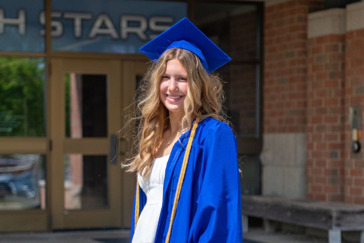 A graduate in a bright blue cap and gown smiles during a graduation ceremony outdoors.