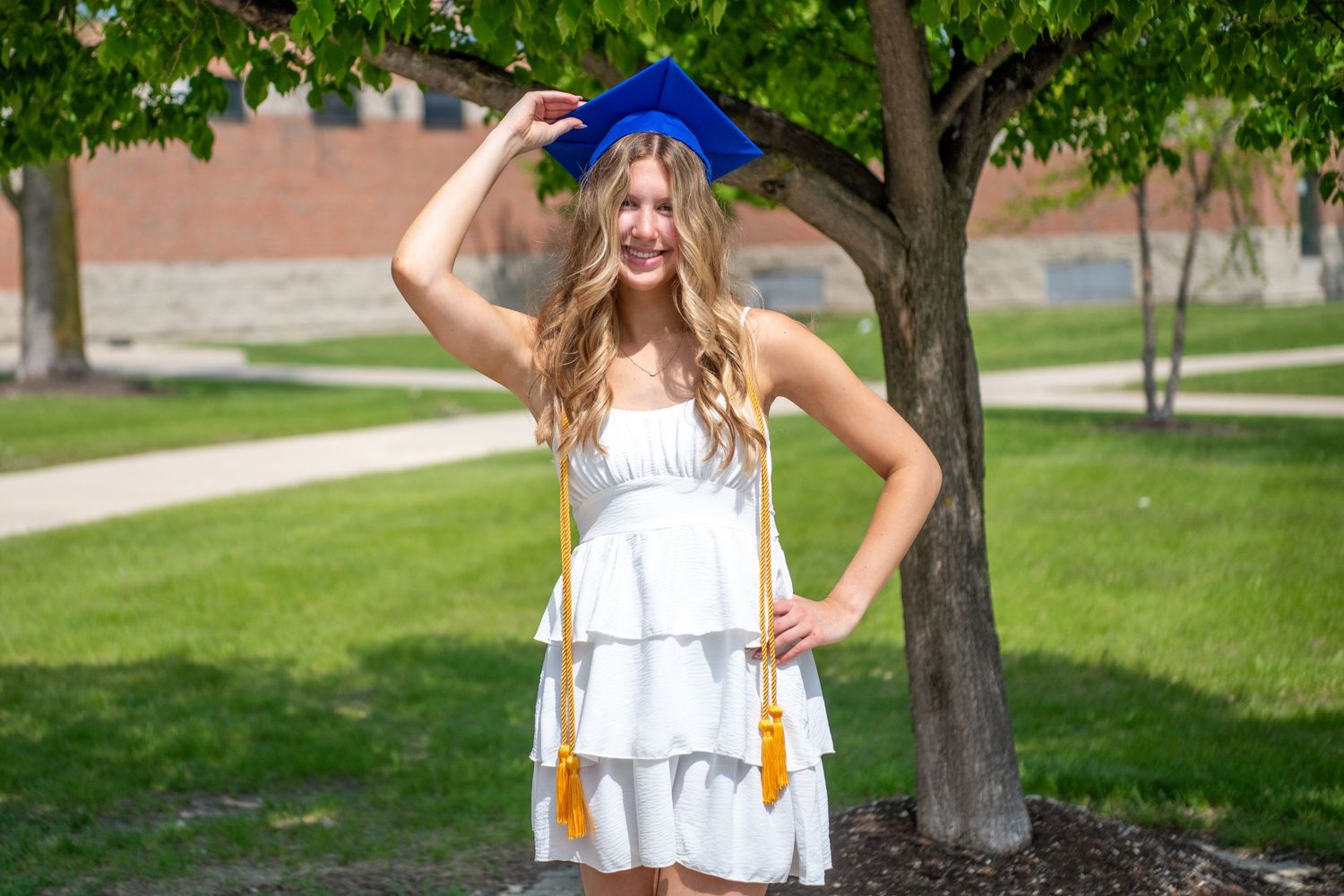 Graduate in white layered dress and blue cap poses outdoors on sunny day near tree on campus lawn.