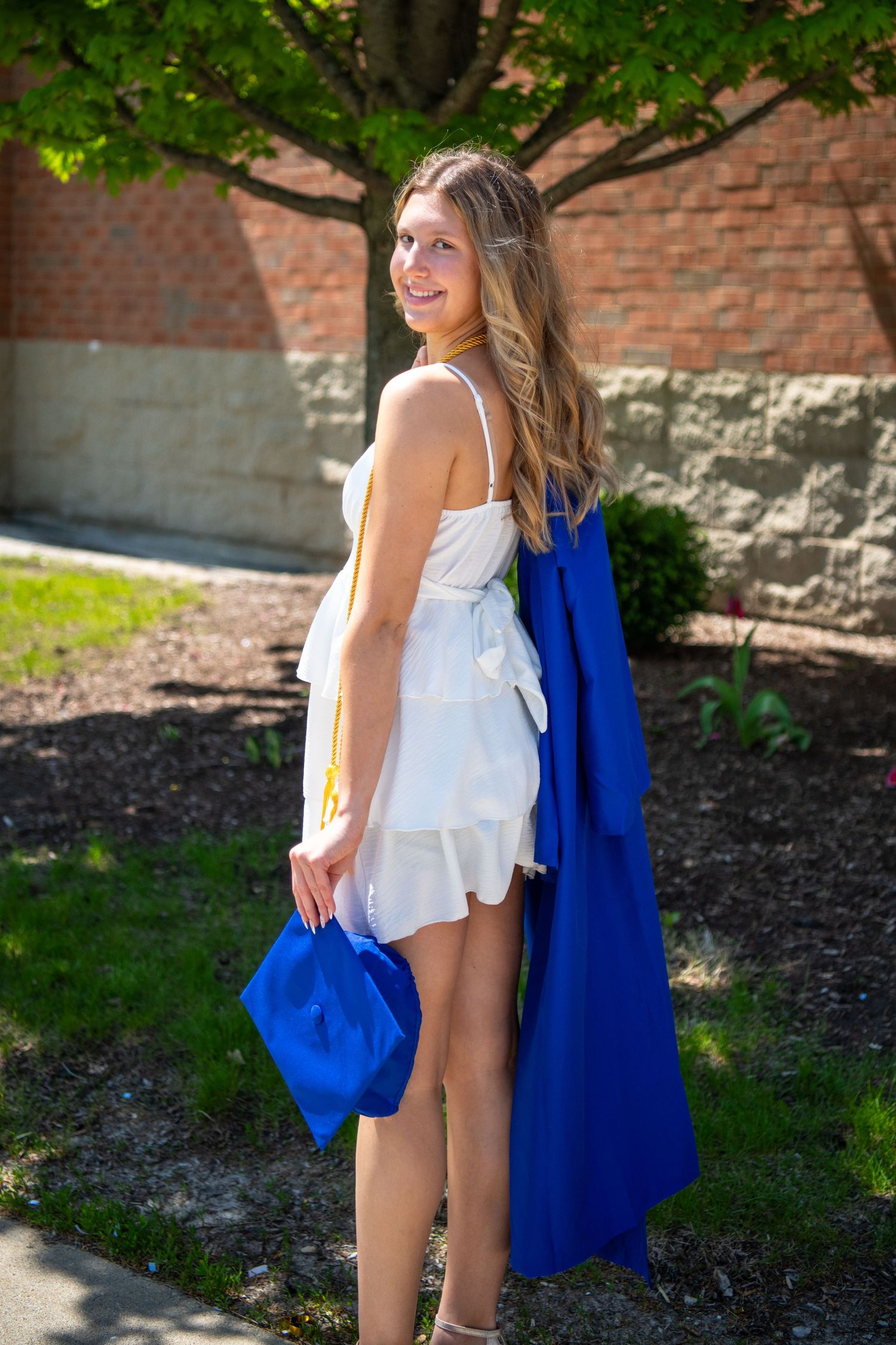 A person wearing a white top and blue graduation gown poses outdoors against a brick wall in natural lighting.